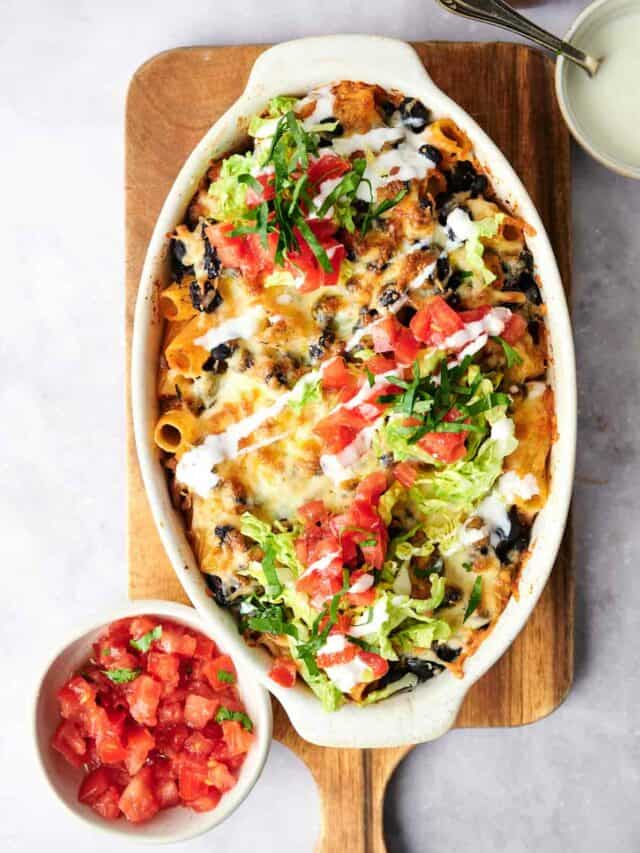 A baked dish of cheesy nachos topped with shredded lettuce, diced tomatoes, and chopped herbs, served on a wooden board with a side bowl of diced tomatoes.