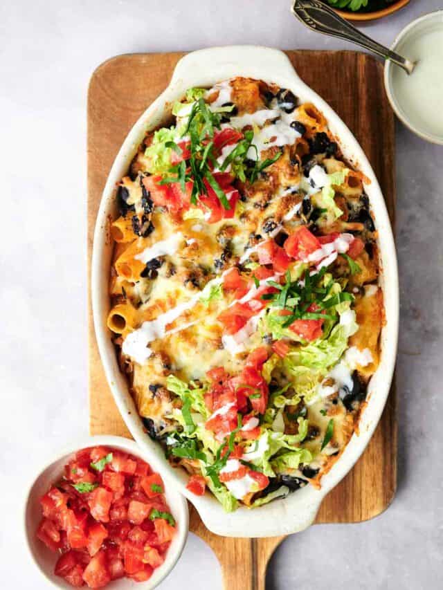 Baked pasta dish topped with melted cheese, fresh tomatoes, and shredded lettuce, served on a wooden board with a side of diced tomatoes.