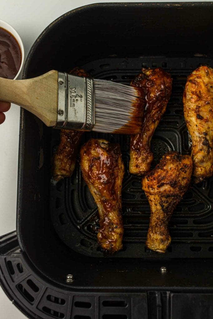 Chicken wings sizzling in an air fryer pan with someone brushing sauce on it.