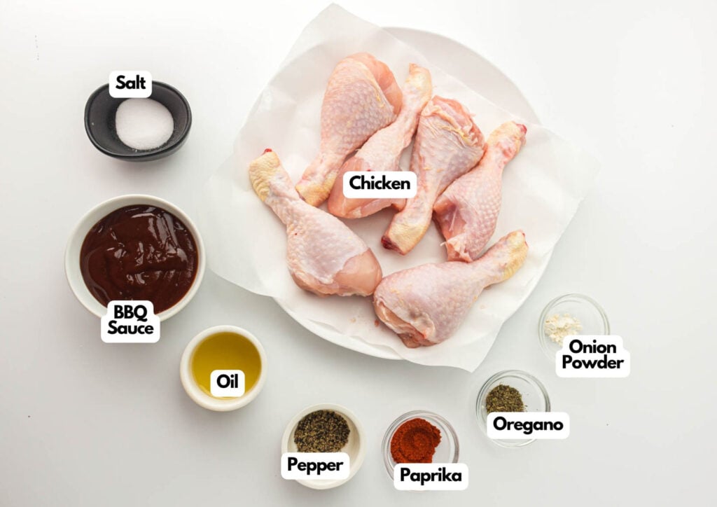 A plate of raw chicken drumsticks surrounded by bowls of salt, BBQ sauce, oil, pepper, paprika, oregano, and onion powder&mdash;everything you need to make flavorful air fryer BBQ chicken drumsticks.