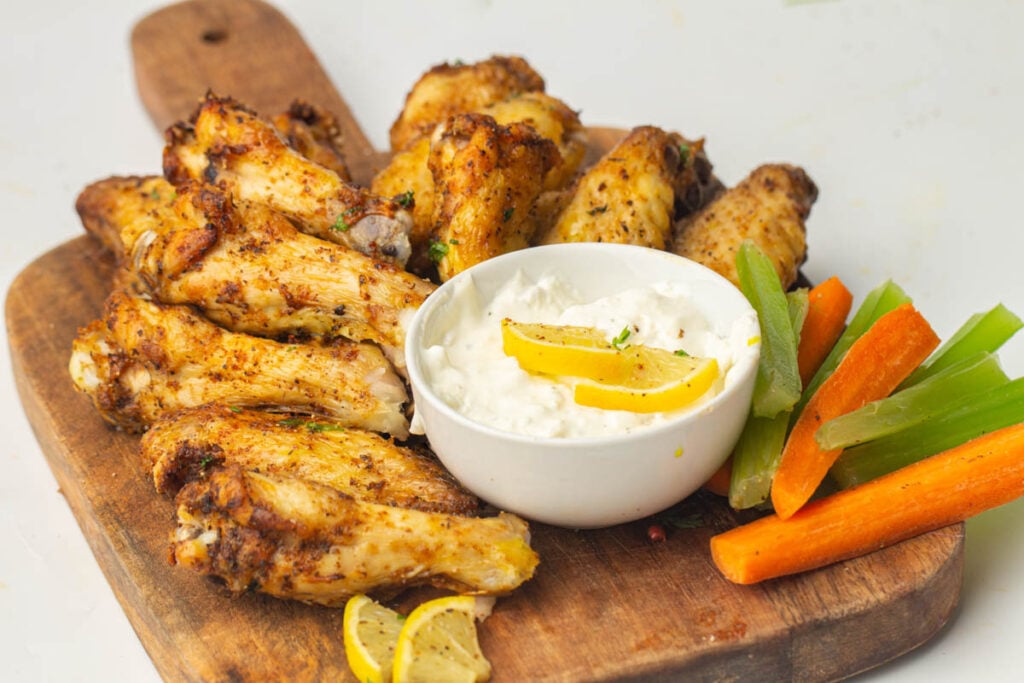 A wooden board with crispy air fryer chicken wings, a bowl of white dipping sauce garnished with lemon, and assorted celery and carrot sticks.