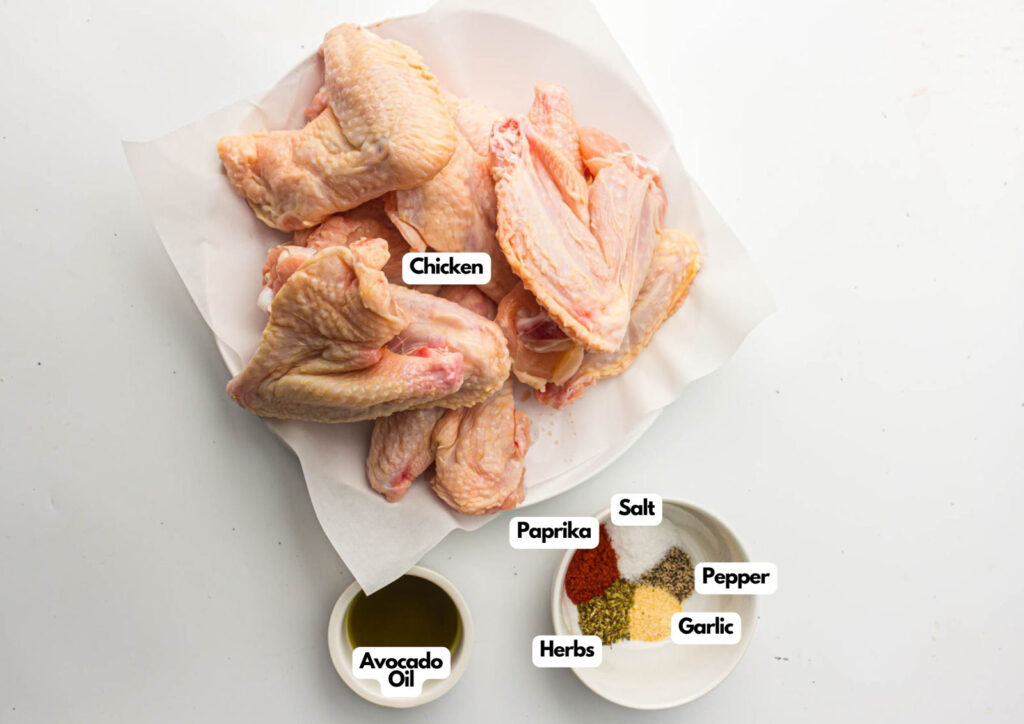 Raw chicken wings on a plate with small bowls of avocado oil, paprika, herbs, salt, pepper, and garlic set beside them—perfect for making flavorful air fryer chicken wings on a white surface.