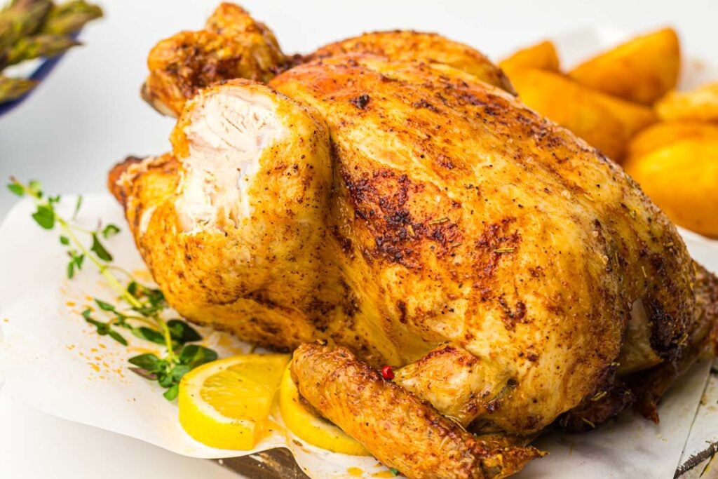 Air fryer whole chicken seasoned with herbs, served on parchment paper with lemon slices and potato wedges in the background.