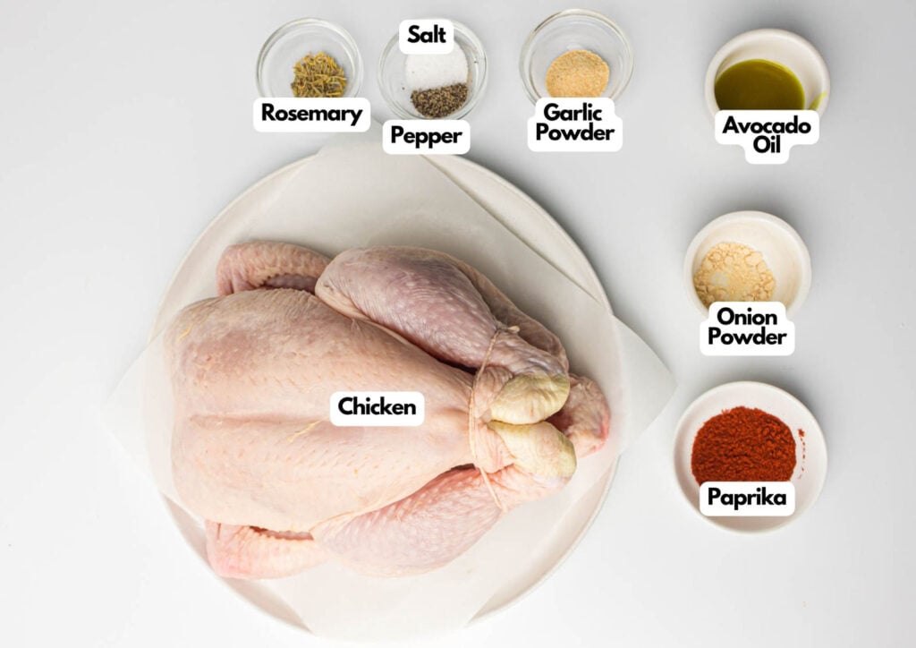 Whole raw chicken on a plate surrounded by labeled bowls of rosemary, pepper, salt, garlic powder, avocado oil, onion powder, and paprika—perfect for making a flavorful air fryer whole chicken.
