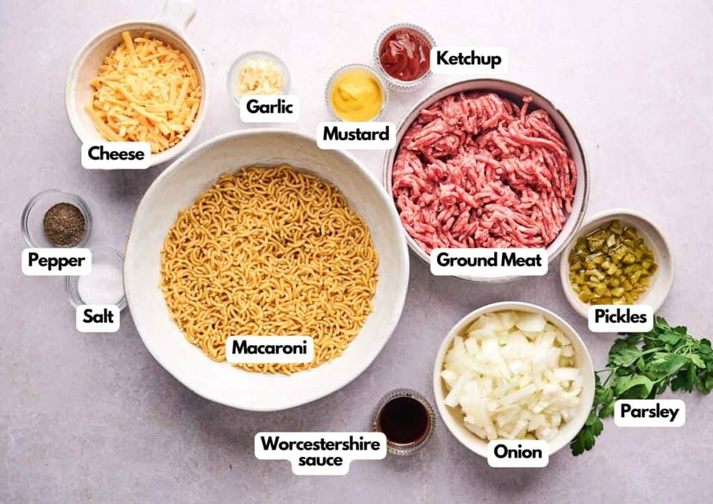 A variety of labeled ingredients for a cheeseburger casserole are arranged in bowls, including ground meat, macaroni, cheese, onion, pickles, ketchup, mustard, and seasonings.