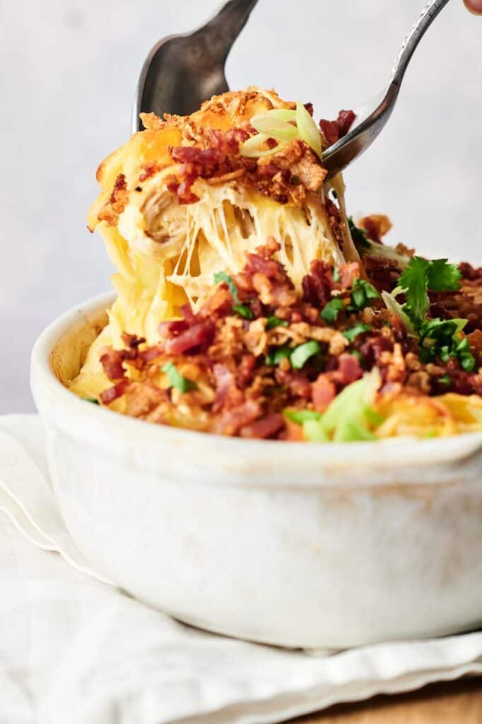 A person holding a fork over a bowl of pasta with bacon and chicken casserole.