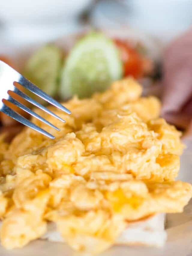 A close-up of a fork piercing scrambled eggs with blurred background of vegetables.