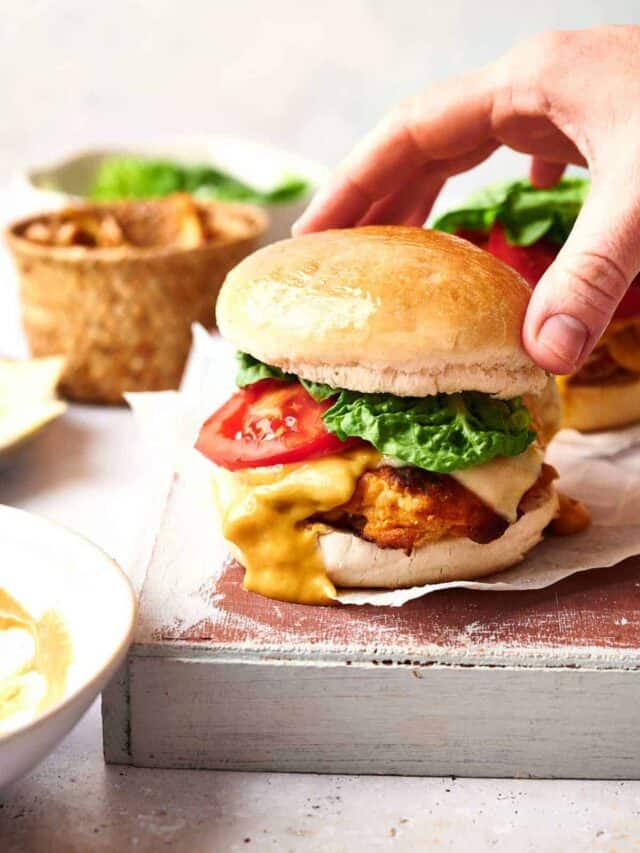 A hand holding a chicken burger with lettuce, tomato, and melted cheese. A bowl of fries and a sauce are in the background.