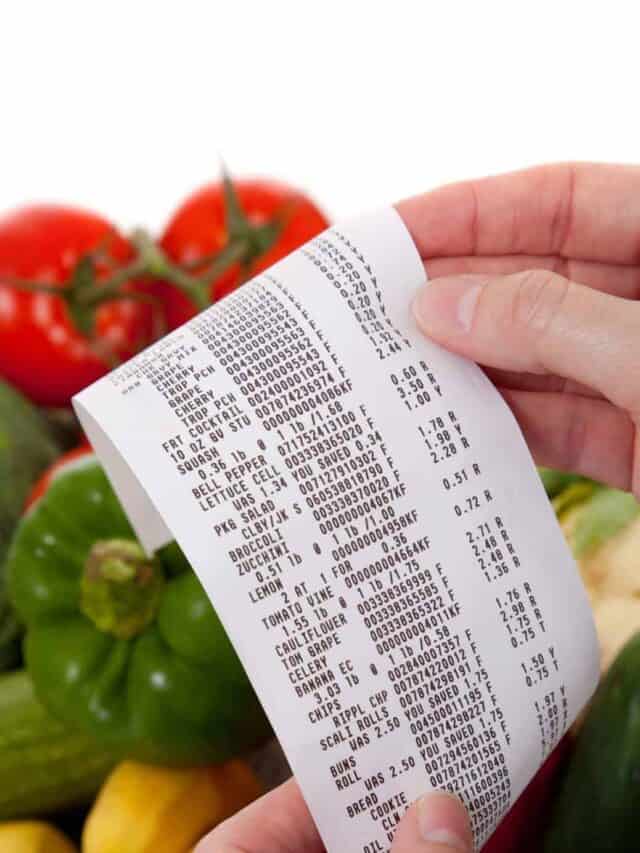 A person holding a grocery receipt in front of colorful fresh vegetables including red tomatoes and green peppers.