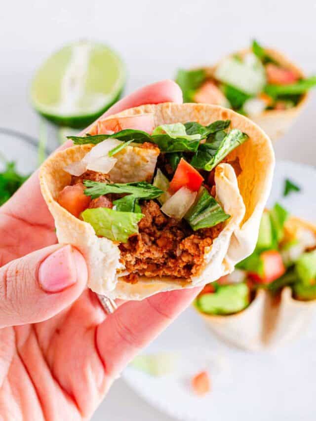 A hand holding a taco filled with ground beef, lettuce, tomatoes, and onions, with lime and more tacos blurred in the background.
