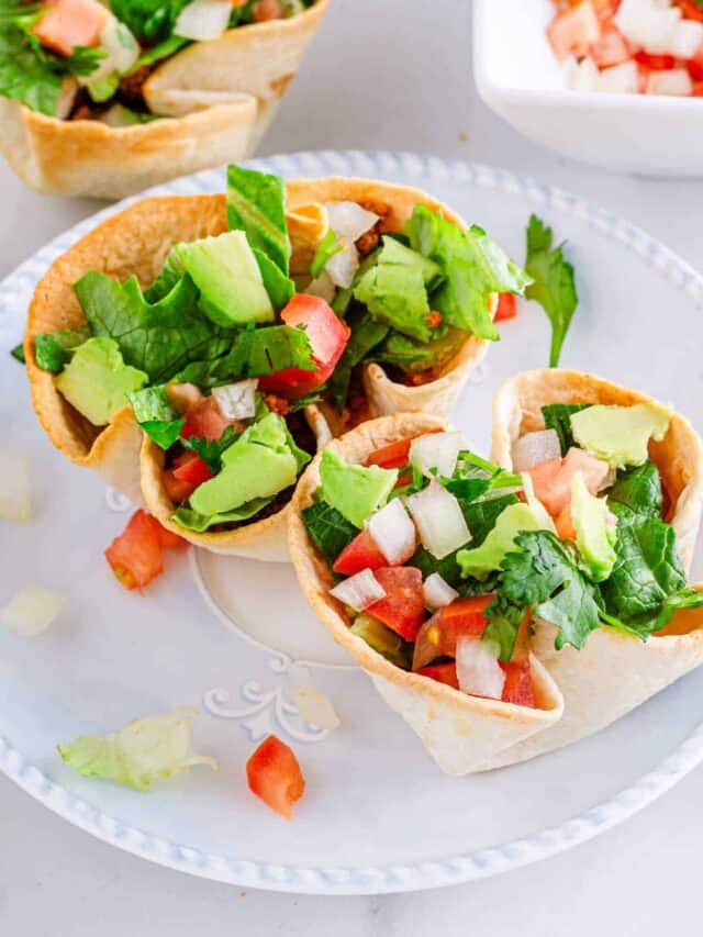 A plate with three taco salad bowls filled with avocado, lettuce, and diced tomatoes.