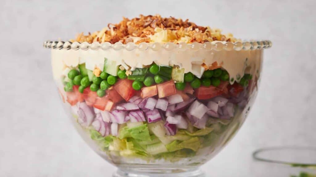 A layered salad presented in a glass bowl featuring lettuce, red onions, tomatoes, peas, cheese, and topped with crispy fried onions.
