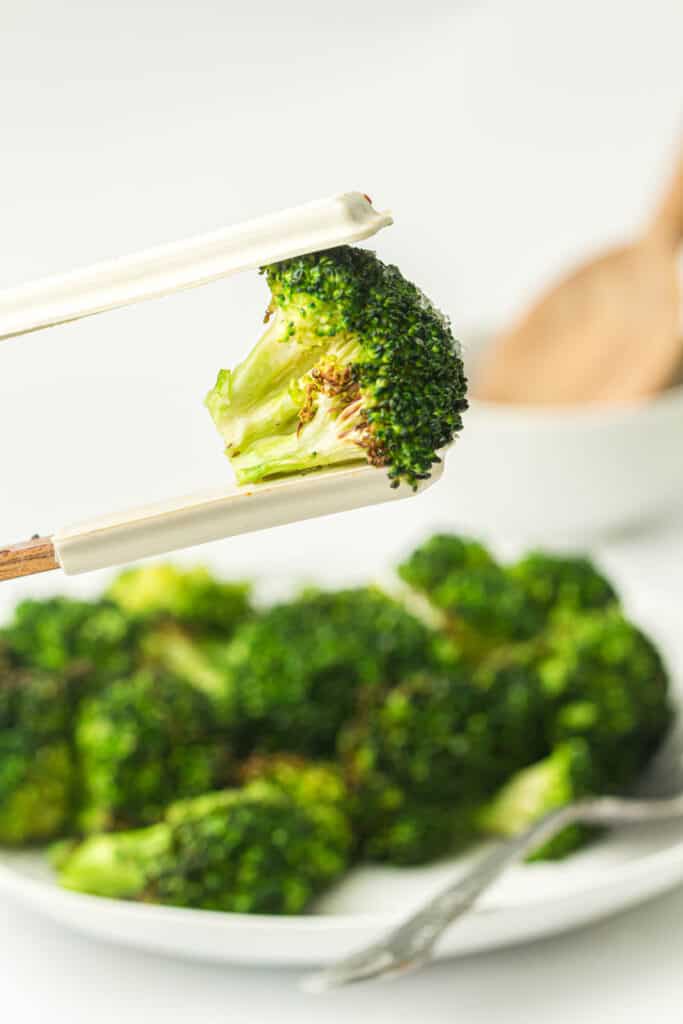 A piece of steamed broccoli held by chopsticks, with a plate of broccoli in the background.