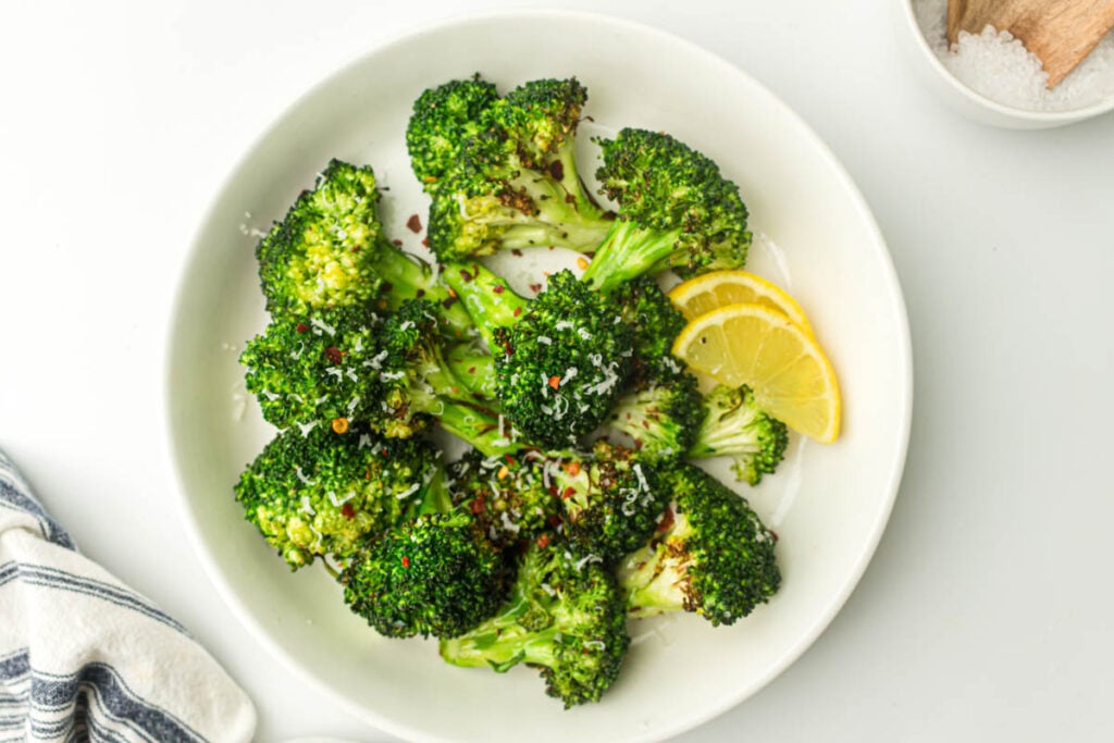 A white plate with air fried broccoli florets, grated cheese, red pepper flakes, and two lemon slices.