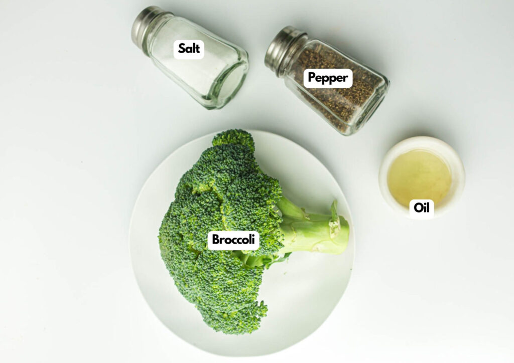 A plate with a head of broccoli, a small bowl of oil, and glass shakers labeled salt and pepper on a white background.