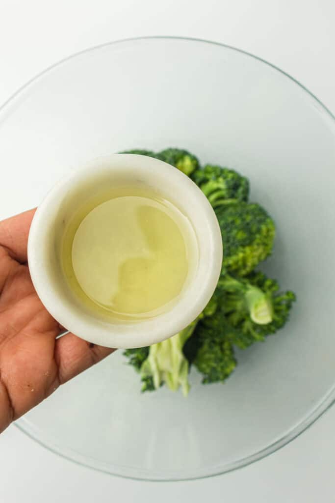 A hand holding a small bowl of avocado oil above a glass bowl containing broccoli florets.