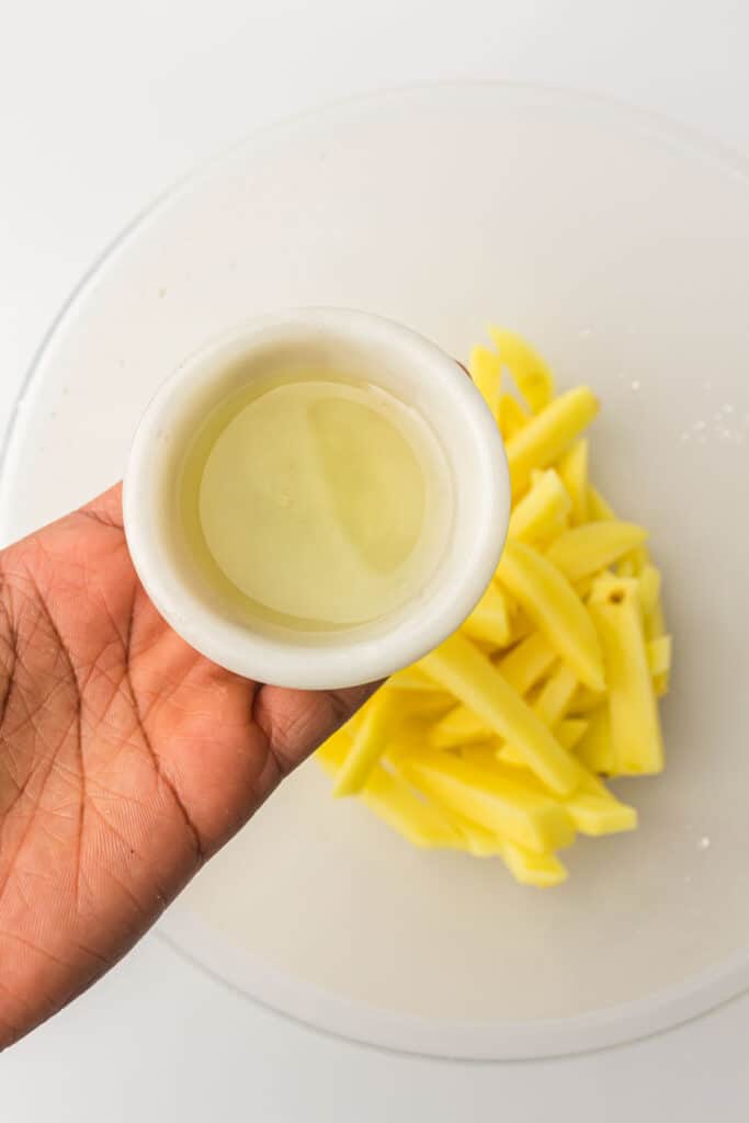 A person holding a small container of oil next to a bowl of sliced raw potatoes.