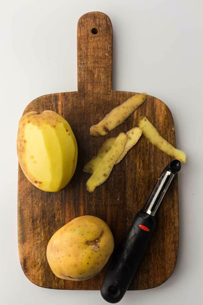 Potatoes with one partially peeled next to a peeler on a wooden cutting board.