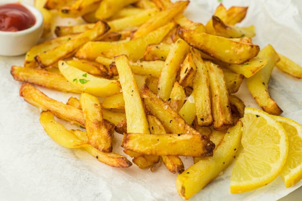 Crispy baked French fries with black pepper, served with lemon wedges and a small dish of ketchup on parchment paper.