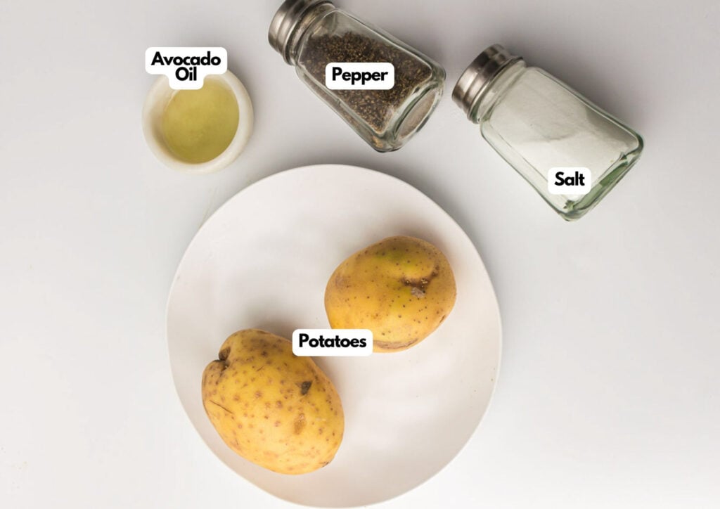 A white plate with two potatoes, next to containers of avocado oil, pepper, and salt, all labeled on a white surface.