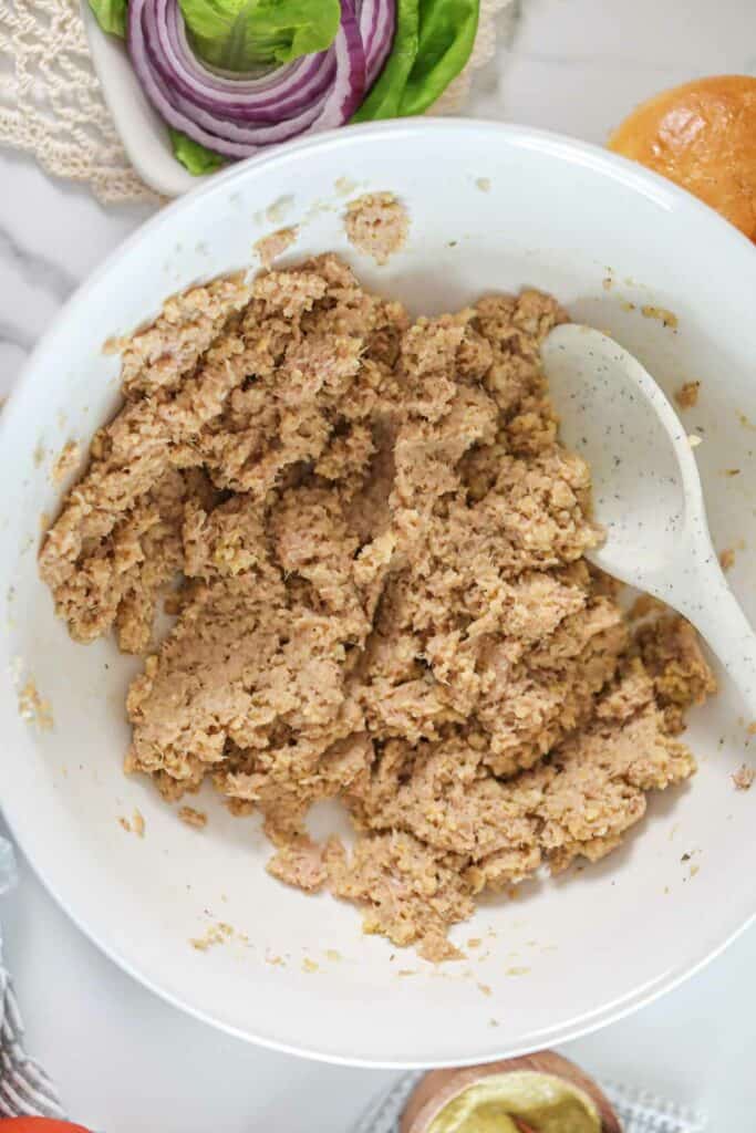 A bowl of ground tuna mixture with a spoon on a kitchen counter, surrounded by various ingredients for air fryer sandwiches.