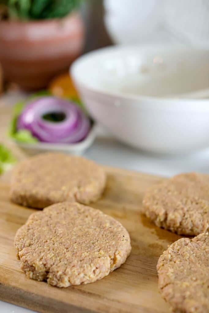 Raw burger patties on a wooden cutting board with a bowl of sauce and sliced vegetables in the background, next to an air fryer.