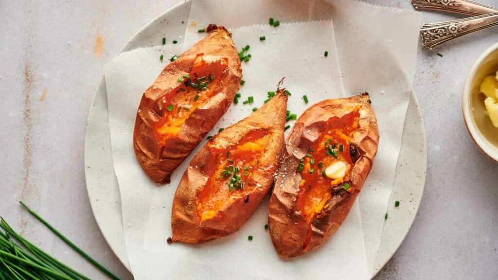 Three baked sweet potatoes topped with butter and garnished with chives, served on a white dish.