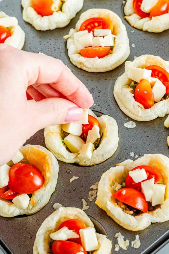 Preparing mini Caprese pesto tarts with tomatoes and cheese on a baking tray.