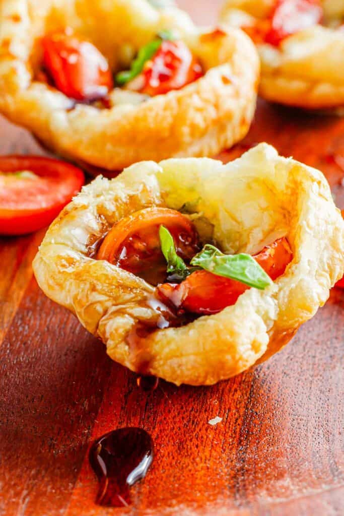 Caprese pesto puff pastry tartlets drizzled with balsamic glaze on a wooden surface.