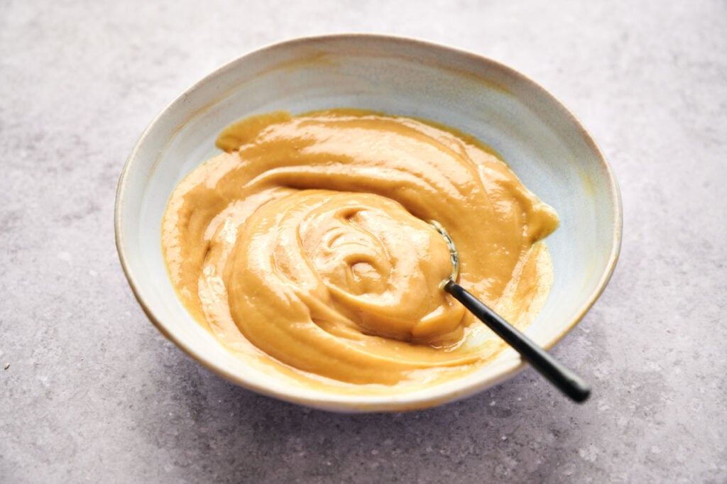 A bowl filled with smooth, light brown Chick-fil-A sauce, with a spoon resting inside, on a light textured surface.