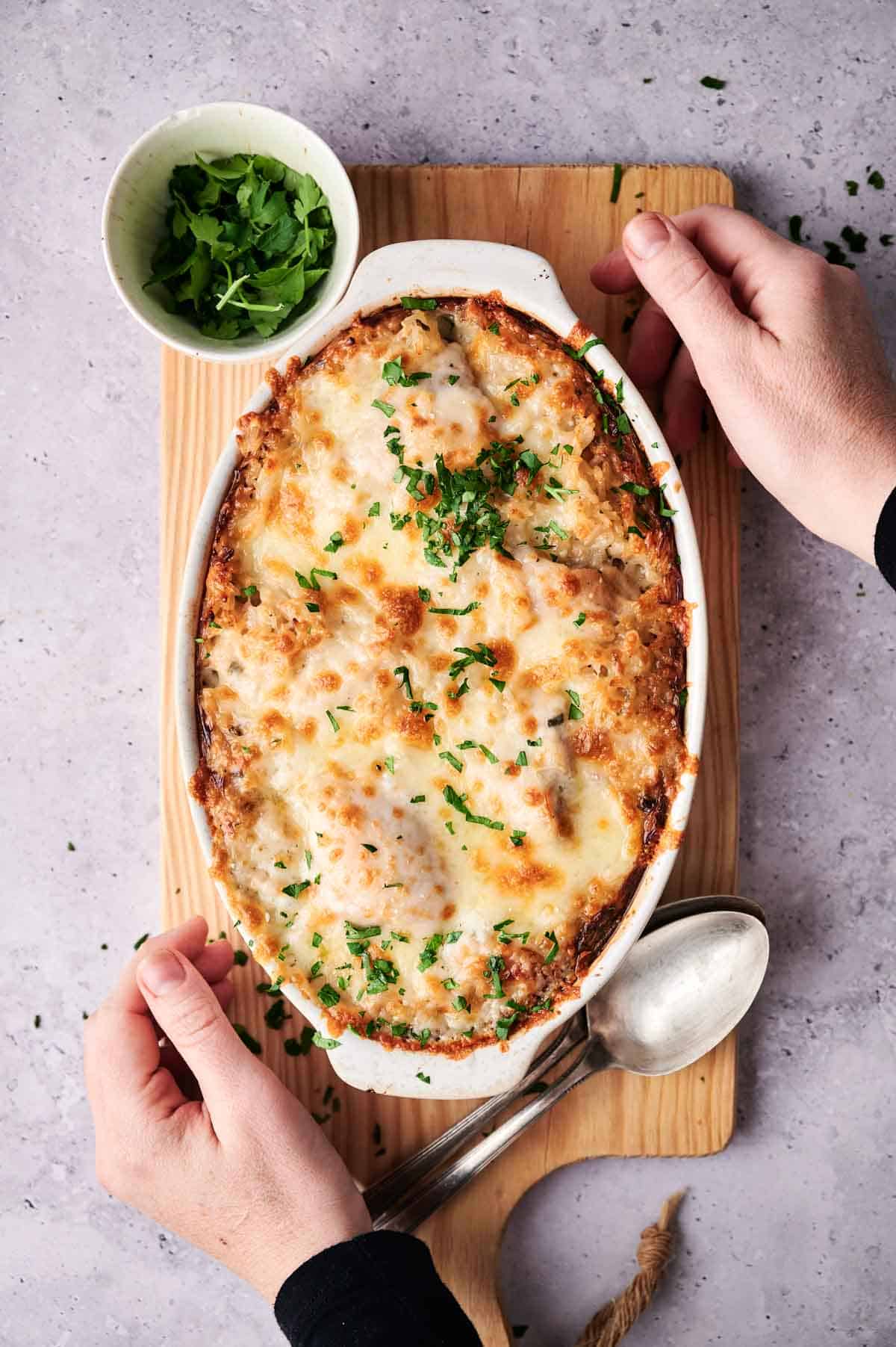 A person serving a freshly baked chicken and rice casserole, garnished with parsley, on a wooden board.