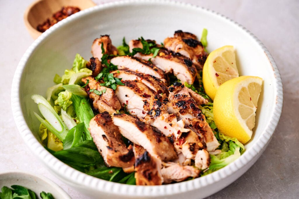 Chipotle grilled chicken, sliced and served on a bed of leafy greens with lemon wedges in a white bowl.