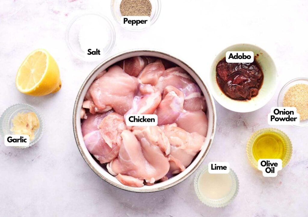 Raw chicken pieces in a bowl surrounded by labeled ingredients—salt, pepper, adobo, onion powder, olive oil, lime, and garlic—ready for a flavorful chipotle grilled chicken marinade on a light surface.