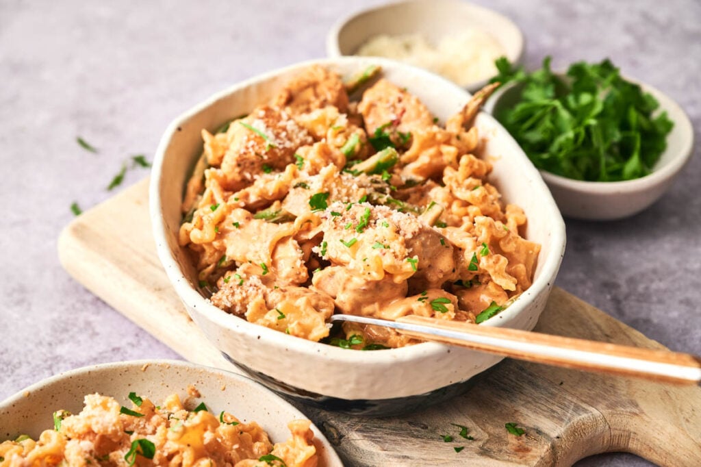 A bowl of creamy Chipotle Pasta with chicken and herbs, topped with grated cheese, sits on a wooden board next to small bowls of grated cheese and fresh chopped parsley.