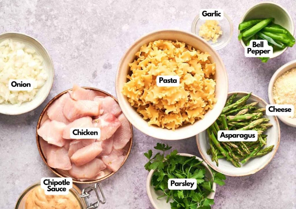 Assorted labeled ingredients for Chipotle Pasta, including chicken, pasta, onion, chipotle sauce, parsley, asparagus, bell pepper, garlic, and cheese—all arranged in separate bowls.