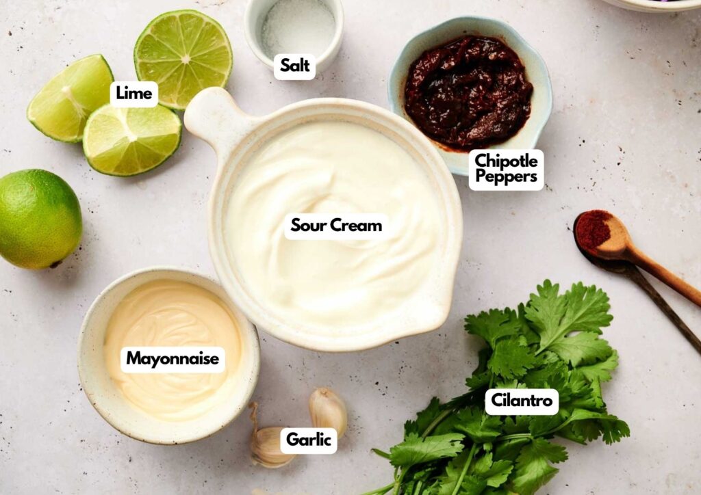 Ingredients for a Chipotle sauce are arranged on a countertop: sour cream, mayonnaise, chipotle peppers, lime, salt, cilantro, garlic, and a spoonful of ground spice.