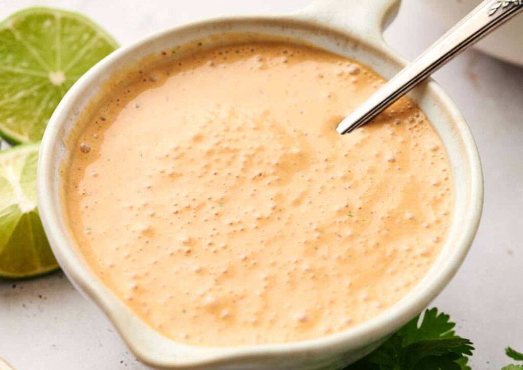 A ceramic bowl filled with creamy chipotle sauce, with a metal spoon inside. Lime slices and cilantro are visible nearby on the table.