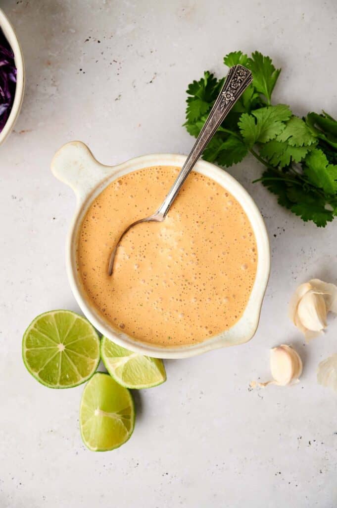 Creamy chipotle sauce in a bowl with a spoon, accompanied by lime wedges, garlic, and fresh cilantro on a light surface.