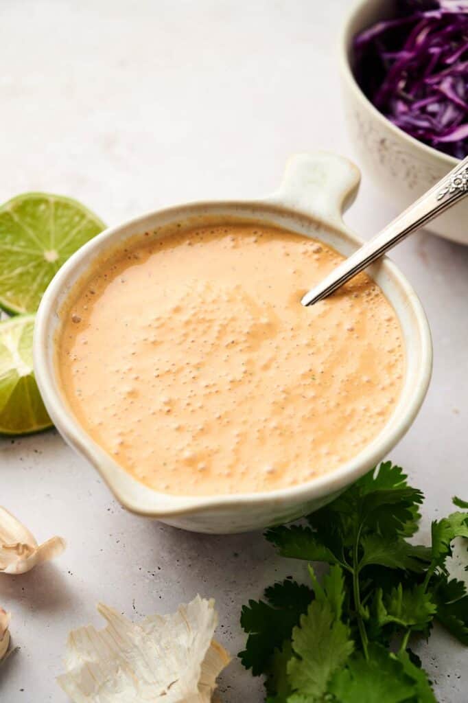 A creamy chipotle sauce in a small bowl, accompanied by lime, garlic, and herbs on a light surface.