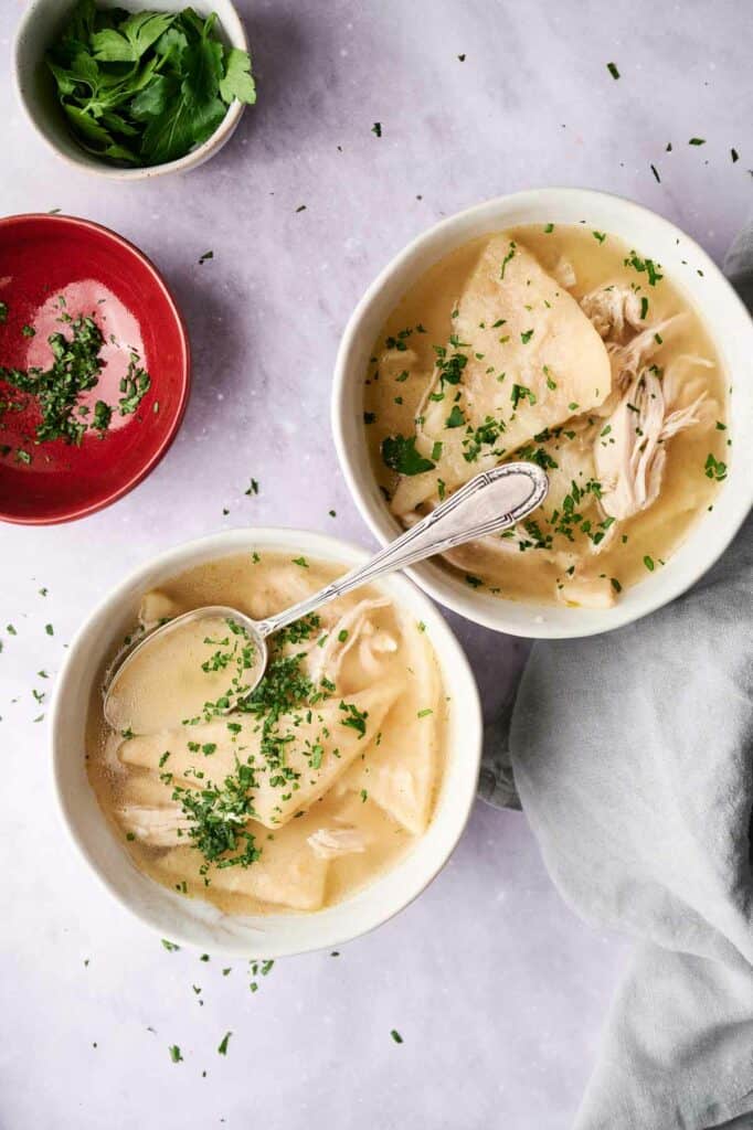 Two bowls of Cracker Barrel Chicken and Dumplings soup garnished with parsley, served on a light surface with a spoon in one bowl and a small bowl of parsley on the side.