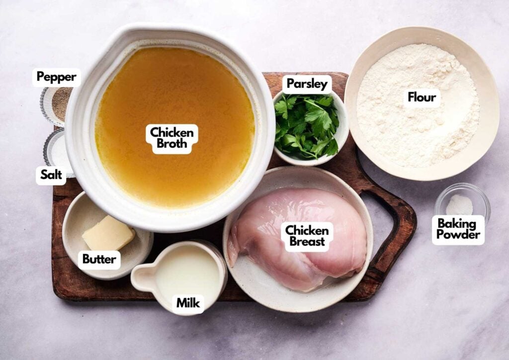 Ingredients for a Cracker Barrel Chicken and Dumplings recipe arranged on a wooden board, including chicken broth, chicken breast, milk, butter, salt, pepper, parsley, flour, and baking powder&mdash;all clearly labeled.