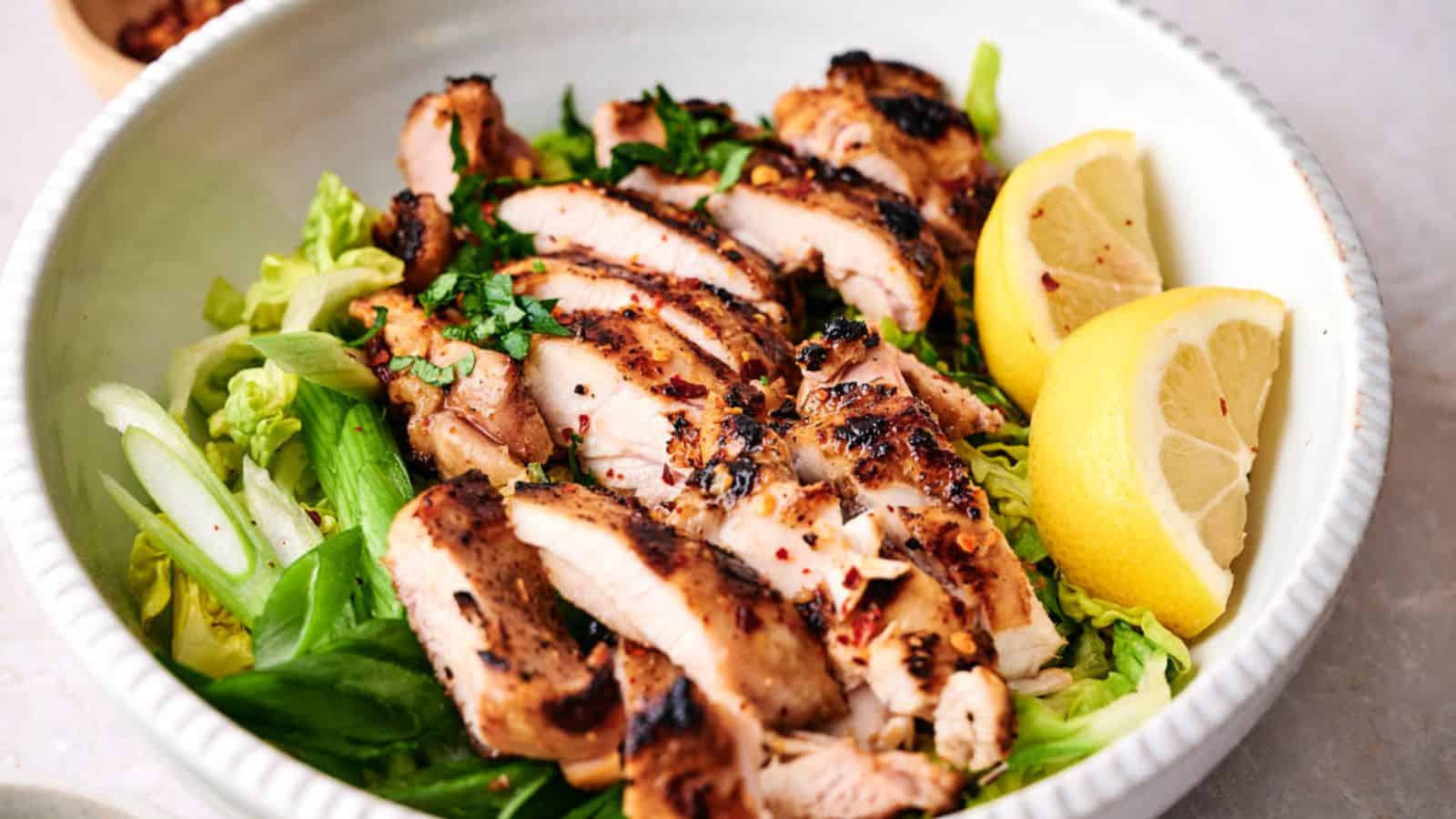 Grilled chicken breast slices served over a bed of greens with lemon wedges on the side.