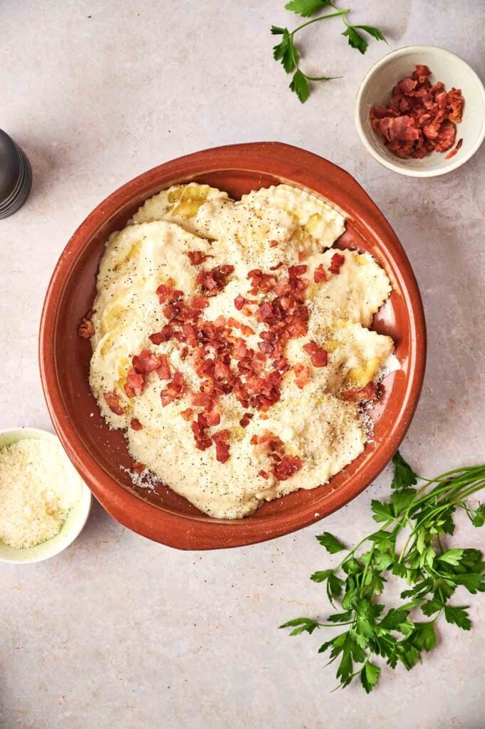 A brown dish of Olive Garden Ravioli Carbonara topped with creamy white sauce, crumbled bacon, and black pepper, surrounded by bowls of bacon, grated cheese, and fresh parsley on a light surface.