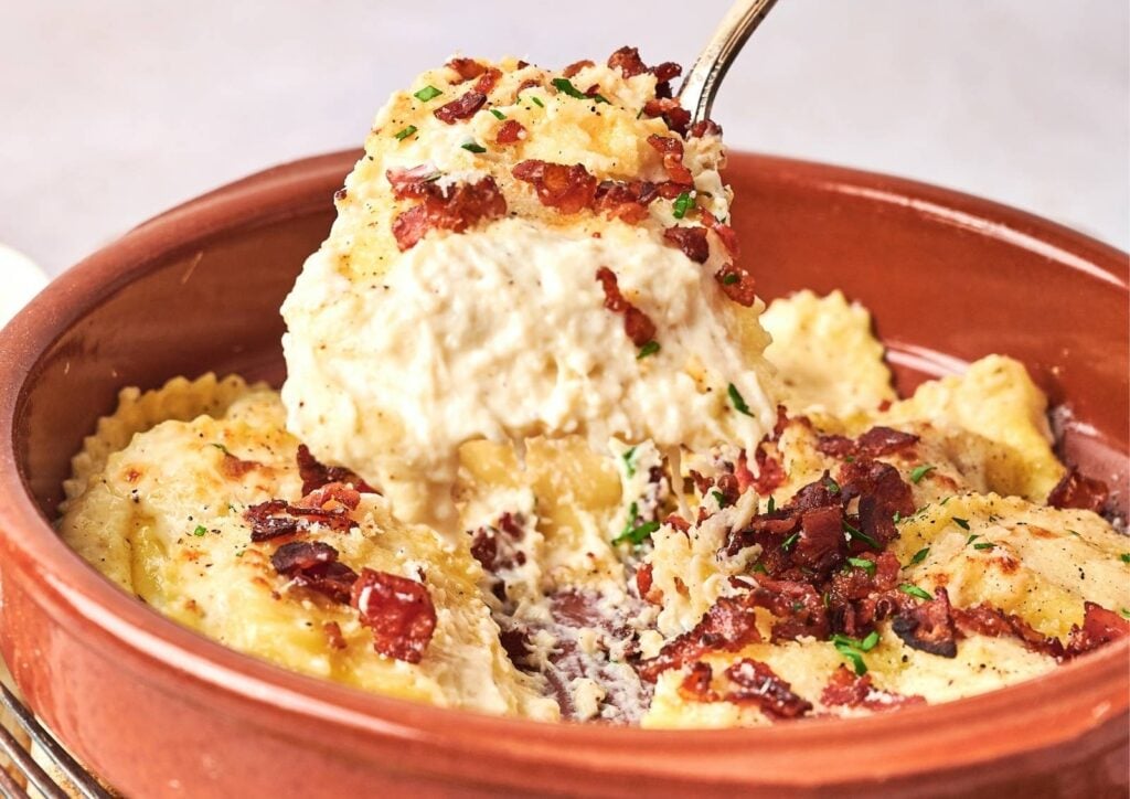 A dish of ravioli topped with creamy white sauce, crispy bacon bits, and chopped herbs, reminiscent of Olive Garden Ravioli Carbonara, served in a brown ceramic bowl with a metal spoon.