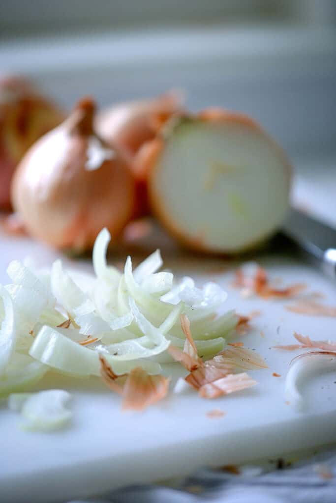 Sliced onions on a cutting board with a knife and whole onions in the background.