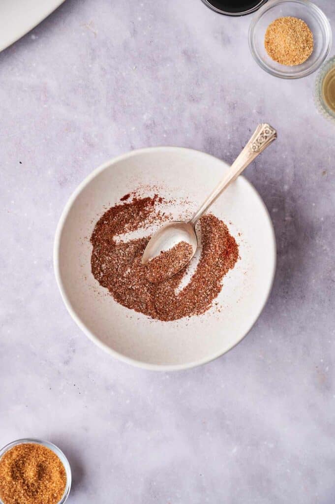 A bowl of ground spices with a spoon, on a kitchen counter alongside other spice containers, prepared for seasoning oven-baked ribs.