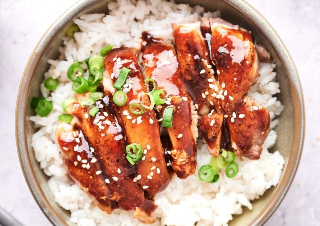 A bowl of white rice topped with sliced Panda Express Teriyaki Chicken, glazed to perfection and garnished with sesame seeds and chopped green onions.