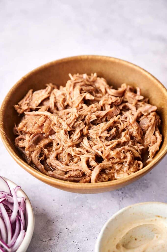 A bowl of Slow Cooker Pulled Pork with sliced onions and a sauce on the side.