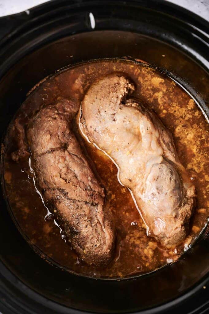 Two pieces of pulled pork cooking in a dark slow cooker.
