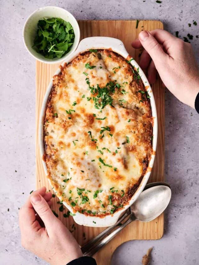 A person holding a baking dish filled with a baked cheesy casserole next to a small bowl of chopped herbs on a wooden board.
